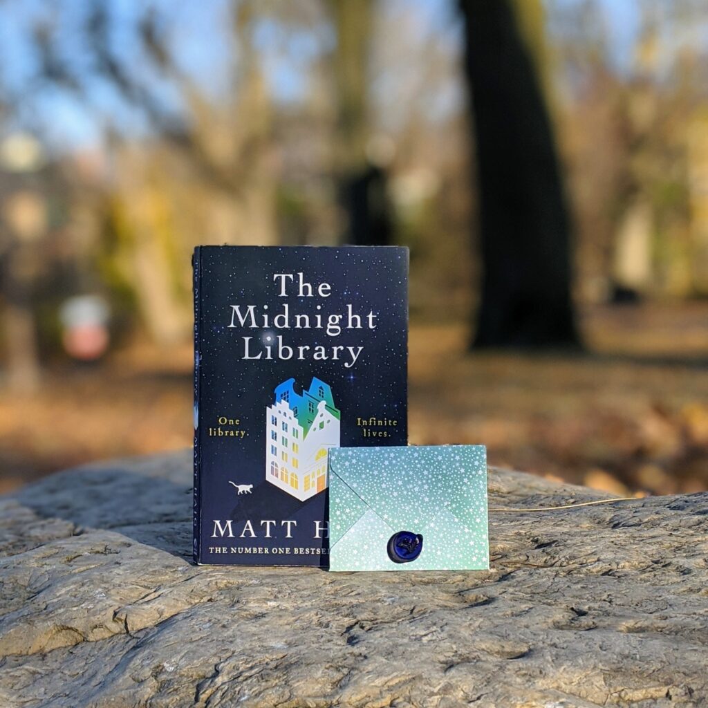 A book and small envelope stand on a rock. The blurred background is of a park with leaves on the ground.